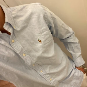 Ljusblå Skjorta från Polo Ralph Lauren (Slim Fit) - Klassisk ljusblå skjorta från Polo Ralph Lauren med broderad logga på bröstet. Skjortan har lång ärm, knappar framtill och button-down krage. Perfekt för en stilren och tidlös look. Storlek Medium Slim Fit. Snabb Leverans!