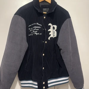 Svart och grå bomberjacka från Bershka - Säljer en snygg bomberjacka i svart och grå manchester från Bershka. Jackan har broderad text och ett stort B på bröstet, knappar framtill och randiga muddar i blått, vitt och grått. Perfekt för dig som gillar streetstyle.