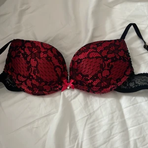 Röd och svart spets-bh från No Secret - Säljer en snygg röd bh med svart spets från No Secret. Bh:n har en liten rosett med en glittrig detalj framtill och justerbara axelband. Sidorna är dekorerade med blommig spets och kuporna har ett vackert mönster.💕