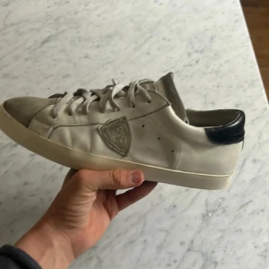 Philip Models  - Säljer ett par vita Philip Models med grå tåparti och svart häl. Skorna har snörning och en platt sula. Sidan pryds av en sköldformad detalj. Perfekta för en avslappnad stil. Lite slitna ✌️ Rabatt om man följer ✅