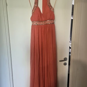 Korallfärgad långklänning från Cotton Club - Säljer en korallfärgad långklänning från Cotton Club med v-ringning och breda axelband. Klänningen har glittriga detaljer med paljetter vid midjan och axelbanden. Perfekt för bal eller festliga tillfällen.