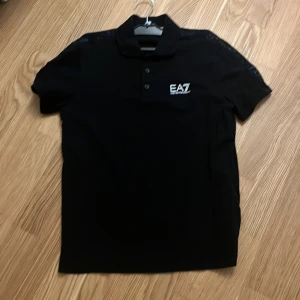 Svart piké t-shirt från Emporio Armani EA7 - Säljer en svart piké t-shirt från Emporio Armani EA7 med klassisk krage och knappar framtill. T-shirten har korta ärmar och EA7-logga på bröstet samt diskret logga på axeln. Perfekt för en stilren look.