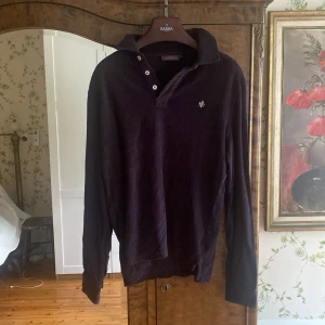 Svart pikétröja från Morris - Klassisk svart långärmad pikétröja från Morris med broderad logga på bröstet. Tröjan har krage, knappar vid halsen och manschettknappar vid ärmsluten. Perfekt för en stilren och avslappnad look.