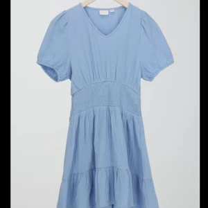 Ljusblå kortklänning från Gina Tricot - Supersöt ljusblå klänning från Gina Tricot med v-ringning och korta puffärmar. Klänningen har smock i midjan och volangdetalj nedtill. Perfekt för sommarens alla tillfällen!