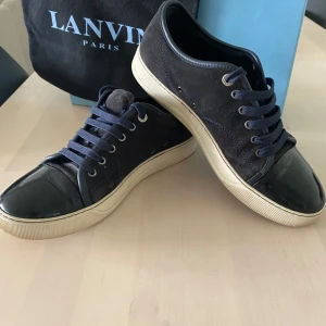 Mörkblåa sneakers från Lanvin - Snygga svarta sneakers från Lanvin med ovandel i mocka och lackad tå. Skorna har vita gummisulor, mörkblå snörning och metallöglor. Klassisk och stilren design med rund tå och platt sula. Storlek 42 - 42/5. 