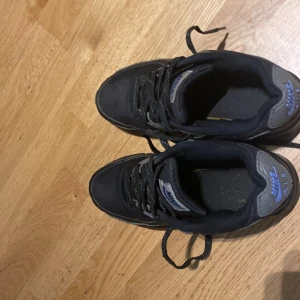 Svarta Nike Air Max sneakers med blå detaljer - Säljer ett par svarta Nike Air Max sneakers med blå swoosh och detaljer. Skorna har snörning, rund tå och en synlig Air Max-sula på sidan. Perfekta för dig som gillar klassisk streetstil och vill ha bekväma sneakers till vardags.