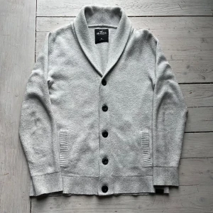 Ljusgrå kofta  - Säljer en stilren ljusgrå kofta med stora svarta knappar och bred krage. Koftan är stickad i mjukt material och har ribbade muddar samt fickor framtill. Perfekt för lager-på-lager och enkel att matcha.