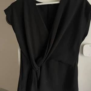 Svart omlottblus - Säljer en svart omlottblus med korta ärmar och djup v-ringning. Blusen har en knytning i midjan som ger en snygg siluett. Perfekt för dig som gillar stilrena och klassiska plagg.