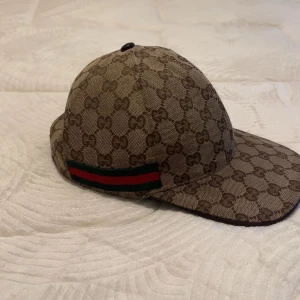 Gucci keps  - Säljer en klassisk Gucci keps i beige med brunt GG-monogram och röd-grön rand på sidan. Kepsen har böjd skärm och justerbart spänne bak i skinn. Perfekt för dig som gillar lyxiga accessoarer.