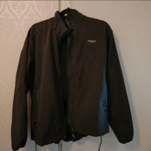 Svart jacka från Pogue med dragkedja - Säljer en svart jacka från Pogue med hög krage och hel dragkedja framtill. Jackan har två sidofickor med dragkedja och justerbar nederkant med snörning. Enkel och stilren design med broderad logga på bröstet.