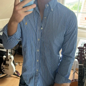 Blåvit randig skjorta från Ralph Lauren - Säljer en klassisk blå och vit randig skjorta från Ralph Lauren med button-down krage och broderad logga på bröstet. Skjortan har långa ärmar och knäppning framtill. Perfekt för en stilren look!