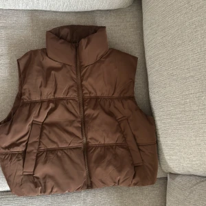 Brun dunväst från H&M - Säljer en brun dunväst från H&M med hög krage och dragkedja framtill. Västen har två snedställda fickor och en enkel, stilren design. Perfekt att slänga över en hoodie eller tröja för extra värme.