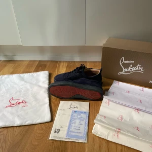 Mörkblå sneakers från Christian Louboutin - Säljer ett par mörkblå sneakers från Christian Louboutin i mocka med klassisk röd sula. Skorna har rund tå, snörning och svarta detaljer. Kommer med originalkartong, dustbag och kvitto.