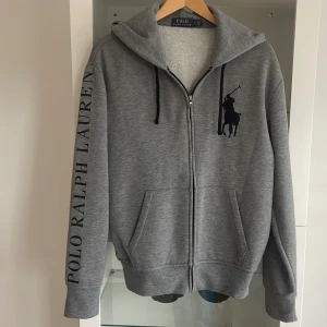 Polo Ralph lauren zip hoodie - Säljer en grå hoodie från Polo Ralph Lauren med dragkedja och stor logga på bröstet. Stora fickor framtill och tryckt text längs ena ärmen. Perfekt för en avslappnad stil.