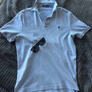 Ljusblå pikétröja från Polo Ralph Lauren - Säljer en klassisk ljusblå pikétröja från Polo Ralph Lauren med krage och två knappar framtill. Tröjan har den ikoniska broderade loggan på bröstet och är perfekt för en stilren look. Storlek S men passar M!