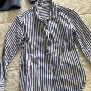 Blåvit randig skjorta från H&M - Säljer en klassisk blåvit randig skjorta från H&M. Skjortan har lång ärm, krage och knäppning framtill. Perfekt för en stilren och avslappnad look. Tillverkad i ett lätt och svalt material. Passar bra till barn 8-9.