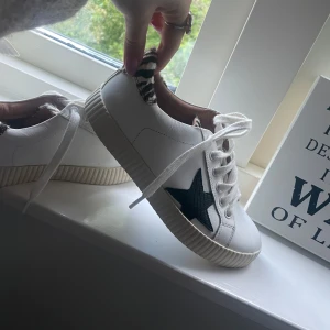 Vita sneakers med svart stjärna och zebramönster - Säljer ett par vita sneakers från  M&S colletion med svart stjärna på sidan och zebramönstrad häl. Skorna har snörning och en tjock, räfflad sula. Perfekta för dig som vill sticka ut med coola detaljer och snygg design.