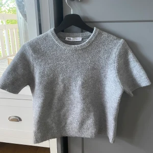 Grå croppad stickad tröja från Zara - Säljer en grå croppad stickad tröja från Zara med korta ärmar och rund halsringning. Tröjan är i storlek M men är väldigt liten i storleken så skulle säga att den sitter som XS-S