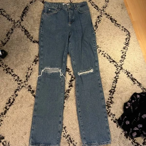 Blå raka jeans med slitna knän från NA-KD - Säljer ett par blå jeans från NA-KD med raka ben och hög midja. Jeansen har slitna detaljer vid båda knäna och klassisk femficksdesign. Storlek 36.