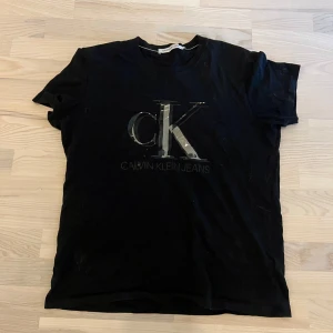 Svart t-shirt från Calvin Klein Jeans - Säljer en svart t-shirt från Calvin Klein Jeans med stort glansigt CK-tryck på bröstet. Klassisk rund halsringning och korta ärmar. Perfekt för en stilren och enkel look.