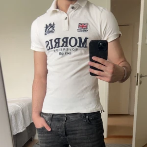 Vit piké t-shirt från Morris - Vit piké t-shirt från Morris med broderad logga och brittisk flagga på bröstet. Klassisk krage med knappar och korta ärmar. Perfekt för en stilren och sportig look. Är i använd men bra skick, finns inga defekter. Är 175cm