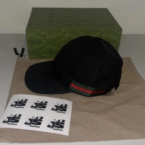 Svart keps från Gucci med grön-röd rand - Snygg svart keps från Gucci med diskret GG-mönster och klassisk grön-röd rand på sidan. Kepsen har böjd skärm och justerbar rem baktill. Perfekt accessoar för en trendig look. Finns i S M L 