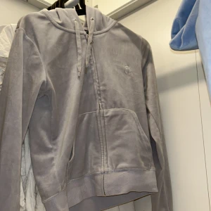 Ljusgrå hoodie från Juicy Couture - Säljer en ljusgrå hoodie från Juicy Couture med dragkedja framtill och fickor. Tröjan har huva med snörning och en liten broderad logga på bröstet. Perfekt för en avslappnad och trendig look. Priset är lågt pga att den tappat metallbitarna på snörerna annars utöver de är de mycket bra skick