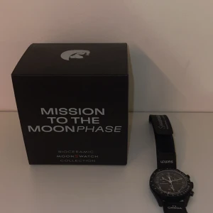 Omega x Swatch MoonSwatch Mission to the Moonphase snoopy  - Säljer en svart Omega x Swatch MoonSwatch Mission to the Moonphase snoopy med biokeramisk boett och svart textilband. Klockan har tydlig tachymeter, kronograf och lysande visare. Levereras i originalförpackning med Snoopy-motiv.