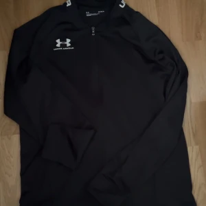 Svart träningströja med dragkedja från Under Armour - Säljer en svart långärmad träningströja från Under Armour med halv dragkedja framtill och logga på bröstet samt text längs ärmen. Perfekt för träning eller sportiga aktiviteter.