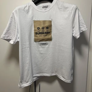 Vit t-shirt från Burberry med beige patch storlek L,använd en gång  - Vit t-shirt från Burberry med en beige fyrkantig patch på bröstet där Burberry-loggan och text syns. Klassisk rund halsringning och korta ärmar. Perfekt för en stilren och trendig look.