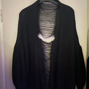 Svart stickad kofta med öppen framsida - Svart stickad kofta med lång ärm och öppen framsida. Koftan har ett unikt, trasigt mönster längs ryggen som ger en edgy look. Perfekt för lager-på-lager och att styla över en enkel topp.