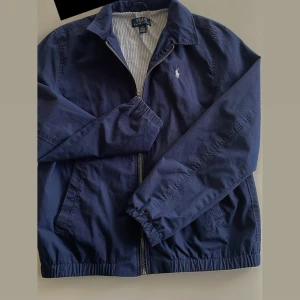 Marinblå vindjacka från Polo Ralph Lauren - Snygg marinblå vindjacka från Polo Ralph Lauren med klassisk krage och dragkedja framtill. Jackan har långa ärmar, elastiska muddar och den ikoniska loggan broderad på bröstet. Perfekt för vår och höst.