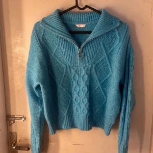 Blå stickad tröja  - Säljer en blå stickad tröja med halv dragkedja och ringdetalj. Tröjan har ett snyggt kabelstickat mönster och hög krage. Perfekt för kyligare dagar och riktigt mysig att ha på sig. Färgen syns bäst på bild 1, det är en knallblå färg perfekt nu till våren 