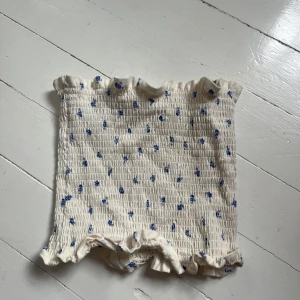 Vit blommig tubtopp med volang - Säljer en söt vit tubtopp med små blå blommor och volangdetaljer upptill och nedtill. Toppen har en smockad struktur som ger en stretchig och bekväm passform. Perfekt för varma dagar! Blir bättre pris om du köper flera. Skriv för fler bilder💙