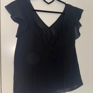 Svart blus med volangärmar köpt i spanien - Säljer en svart blus med v-ringning och fina volangärmar. Blusen har spetsdetaljer vid halsringningen och är tillverkad i ett lätt, tunt material som ger ett elegant intryck.