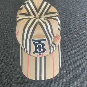 Beige rutig keps från Burberry - Säljer en klassisk beige keps från Burberry med svart, vit och röd rutig design samt broderad TB-logga framtill. Kepsen är tillverkad i bomull och har en normal passform. Perfekt för dig som gillar stilrena accessoarer.