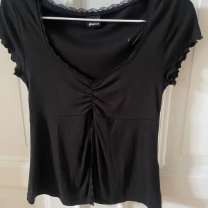 Svart topp med spetsdetaljer från Gina Tricot - Säljer en svart topp från Gina Tricot med korta ärmar och v-ringning. Toppen har fina spetsdetaljer längs halsringningen och framtill, samt rynkade detaljer vid bröstet. Slits på magen (svårt att visa på bild så skriv privat för tydligare bild). Jättefin och använd kanske 1 gång gissar jag på. 