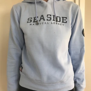 Ljusblå Hoodie Stl 36 - Ljusblå hoodie med trycket "SEASIDE NAUTICAL LEGACY" framtill. Hoodien har dragsko och muddar vid ärmslut och nederkant. Det finns även dekorativa märken på ärm och nertill.  Bara använd en gång, är i nyskick förutom en liten liten fläck fram se bilden. Nypris: 599:-