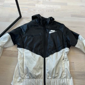 Svart och vit vindjacka från Nike - Nike vindjacka i svart och vit med dragkedja framtill och huva. Jackan har en sportig look med logga på bröstet och praktiska fickor. Perfekt för blåsiga dagar och enkel att matcha till olika outfits.