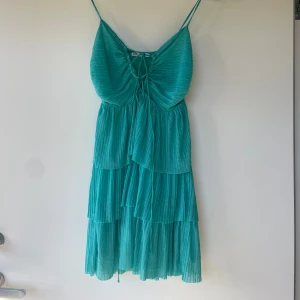 Turkos plisserad klänning från Zara - Säljer en turkos plisserad klänning från Zara med smala axelband och volangdetaljer. Klänningen har omlottkänsla framtill med knytning vid bysten och är tillverkad i ett lätt, luftigt material. Perfekt för varma dagar!
