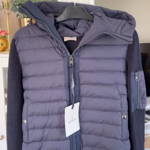 Mörkblå pufferjacka med stickade ärmar från Moncler - Snygg mörkblå pufferjacka från Moncler med stickade ärmar och huva. Jackan har dragkedja framtill, två fickor med tryckknappar och en Moncler-logga på ärmen. Perfekt för kyliga dagar och ger en sportig look.