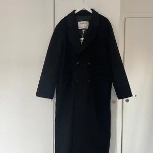 Svart lång kappa från Brixtol Textiles, XL - Säljer en svart lång kappa från Brixtol Textiles i klassisk modell med dubbelknäppning och bred krage. Helt ny med prislapp. 50% ull 50% återvunnet polyester 