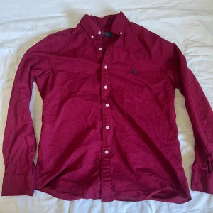Vinröd linneskjorta från Polo Ralph Lauren - Vinröd långärmad skjorta från Polo Ralph Lauren med klassisk krage och knappar framtill. Broderad logga på bröstet i mörkblått. Skjortan är i linne och har en tidlös, normal passform. Perfekt för dig som gillar stilrena plagg med ikonisk touch.