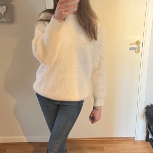 Vit fluffig stickad tröja - Mysig vit stickad tröja med fluffig känsla och rund hals. Tröjan har lång ärm och en avslappnad passform, perfekt för kyligare dagar. Enkel och stilren design som passar till mycket. Aldrig använd då den är för kort i armarna på mig. 