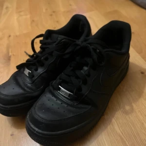 Svarta Nike Air Force 1 sneakers - Säljer ett par klassiska helsvarta Nike Air Force 1 sneakers med svart snörning och låg profil. Skorna har perforerad tå och en platt sula, perfekt för en stilren vardagslook.
