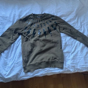 Olivgrön tröja med blixtmönster från Neil Barrett - Säljer en olivgrön tröja från Neil Barrett med ett coolt blixtmönster i svart  över bröstet. Ny pris på denna är runt 2000-3000 mitt pris 699 priset kan diskuteras. Skriv om ni vill ha mer bilder.