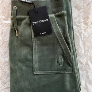 Mossgröna velourbyxor från Juicy Couture - Mossgröna (moss green) Classic Velour Del Ray byxor från Juicy Couture med raka ben, dragsko i midjan och fickor fram. Byxorna har broderad logga på ena fickan och en bakficka med knapp. Fråga för mer info, (pris etc).