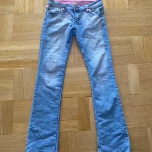 Blå jeansbyxor med rosa detaljer från Revers Jeans - Säljer ett par blå jeansbyxor från Revers Jeans med rosa sömmar och detaljer, samt broderad text på linningen. Byxorna har bootcut-modell och bakfickor med slitna detaljer och rosa kantband.