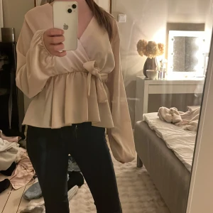 Beige omlottblus med knyt - Säljer en beige omlottblus med lång ärm och knyt i midjan. Blusen har en v-ringad design och volang nedtill 