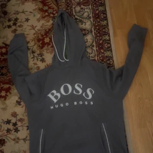 Grå hoodie från Hugo Boss - Säljer en grå hoodie från Hugo Boss med stor vit logga på bröstet. Tröjan har huva med vita snören och en klassisk känguruficka framtill. Perfekt för en avslappnad och stilren look.